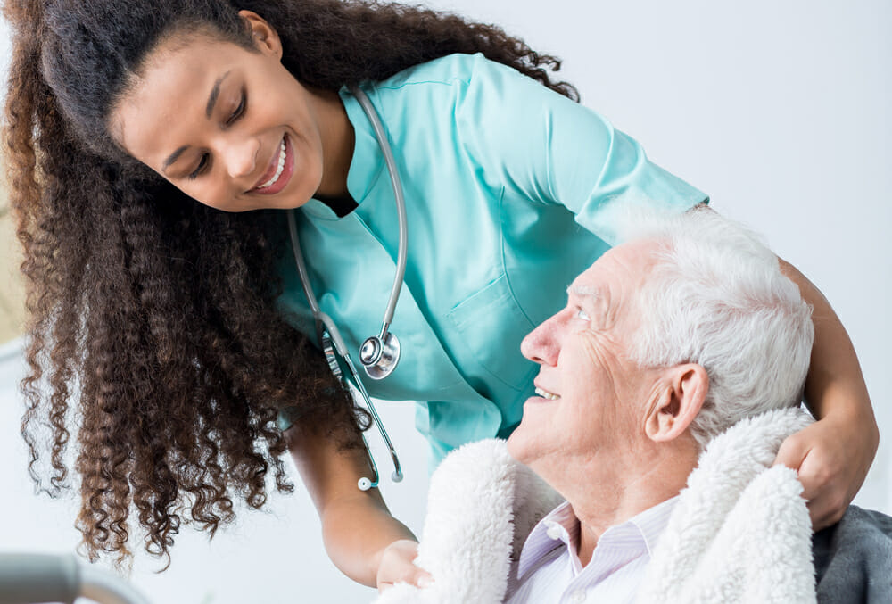 Young female caregiver and elderly man covered with blanket smiling to each other